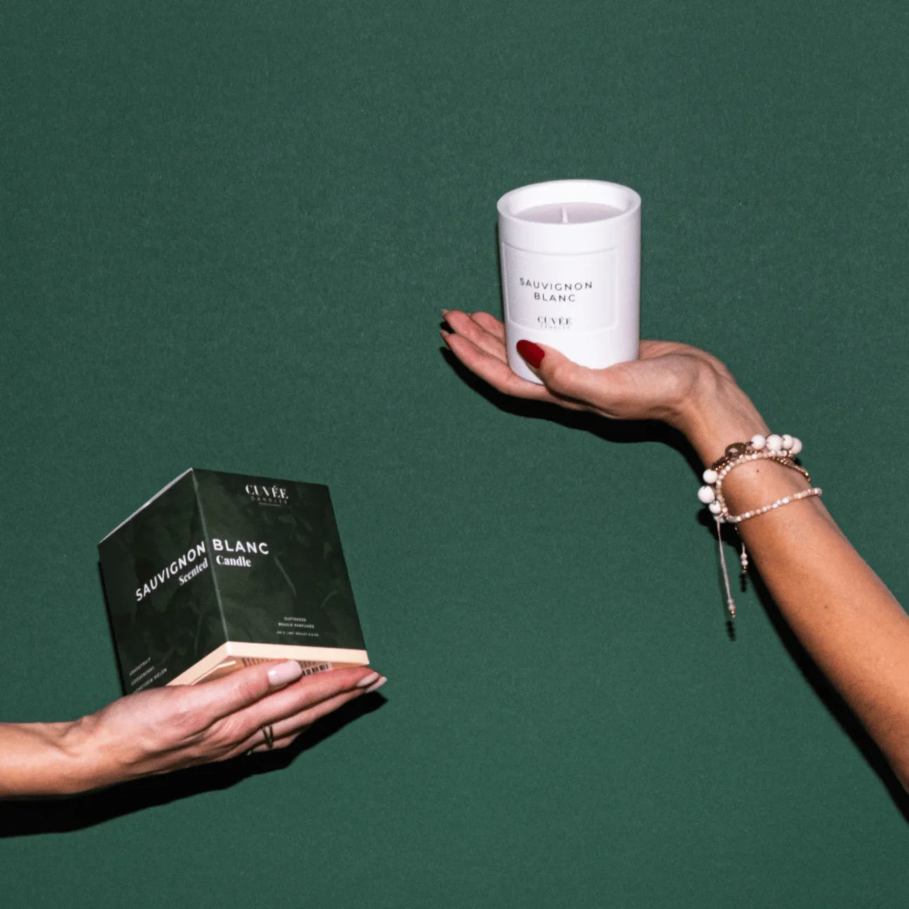 Hands of the two CUVÉE CANDLES founders and sisters Katharina and Miriam holding the box of the Sauvignon Blanc candle and the scented candle in the air against a green background.